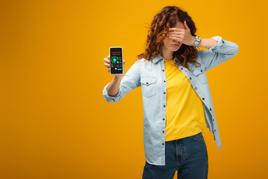 redhead vrouw die ogen en het houden van smartphone met marketing analyse op het scherm op oranje  - Foto, afbeelding