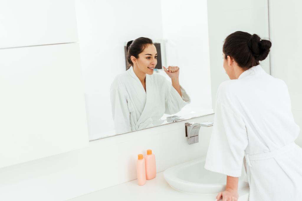 Selektywny fokus wesoły kobieta patrząc na lustro podczas szczotkowania zębów w łazience  - Zdjęcie, obraz