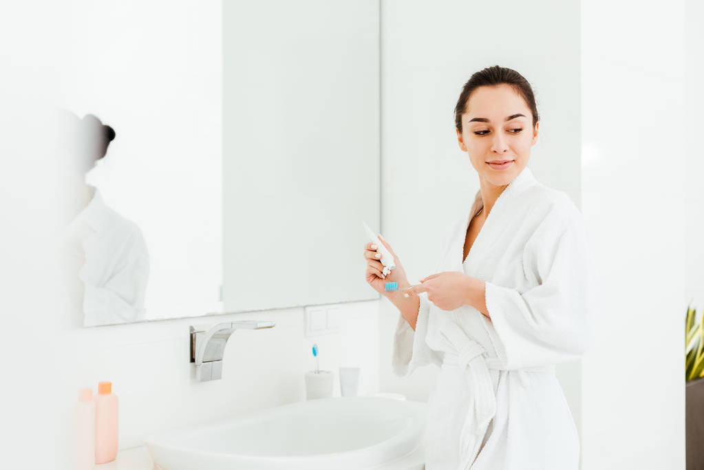 вибірковий фокус брюнетки, що стоїть біля дзеркала, тримаючи зубну щітку і зубну пасту
   - Фото, зображення