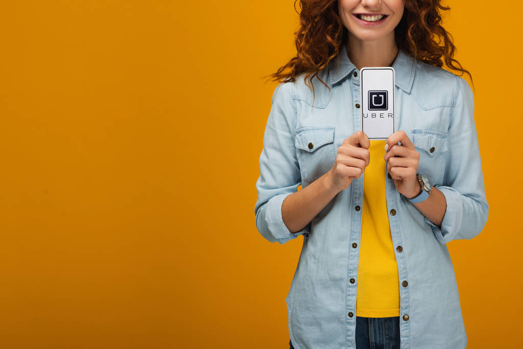 przycięte widok wesoły kręcone kobieta trzyma smartfon z Uber aplikacji na ekranie na pomarańczowo  - Zdjęcie, obraz