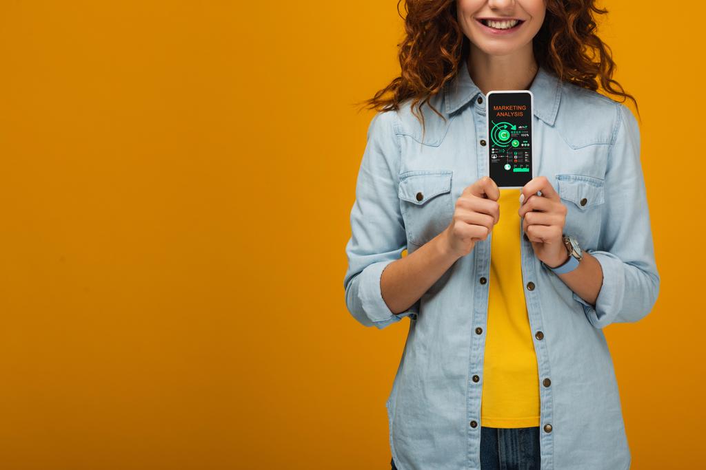 オレンジ色の画面上のマーケティング分析とスマートフォンを保持する陽気な巻き毛の女性のトリミングビュー  - 写真・画像