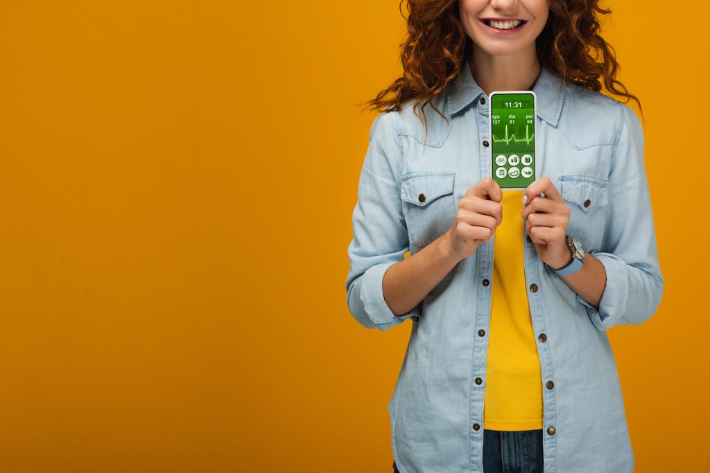 cropped view of cheerful curly woman holding smartphone with e-health app on screen on orange  - Photo, Image