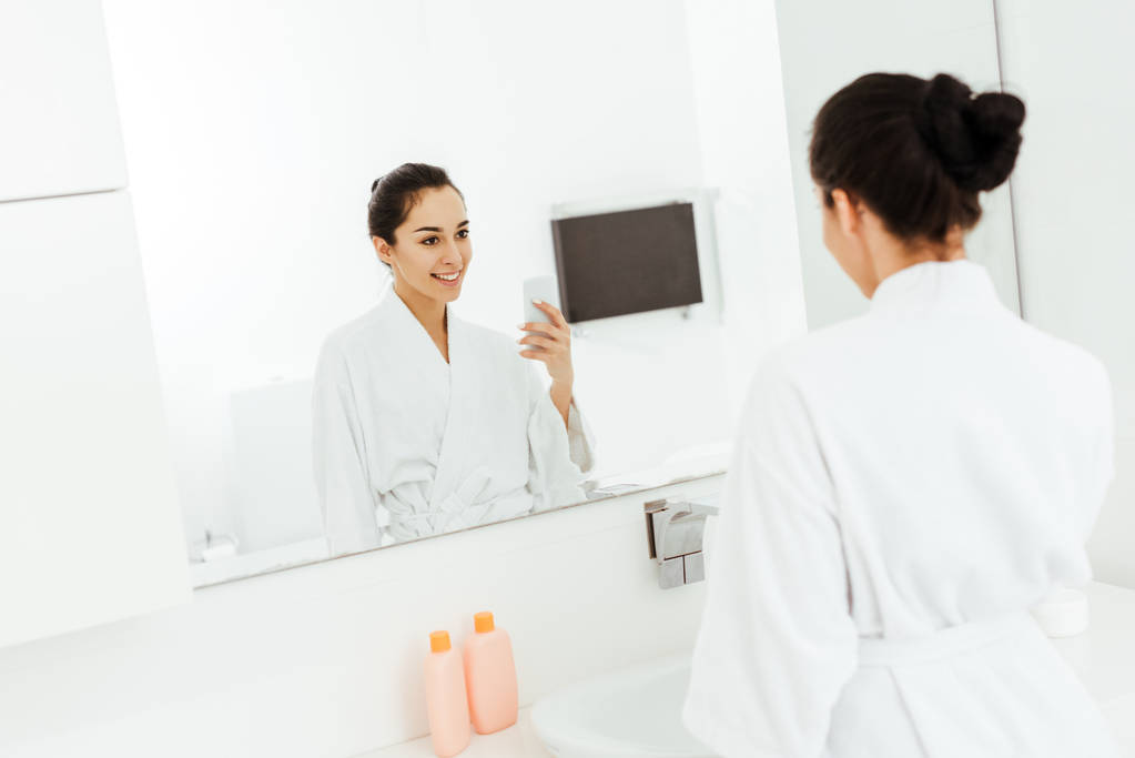 selectieve focus van aantrekkelijke en vrolijke jonge vrouw die foto in de badkamer neemt  - Foto, afbeelding