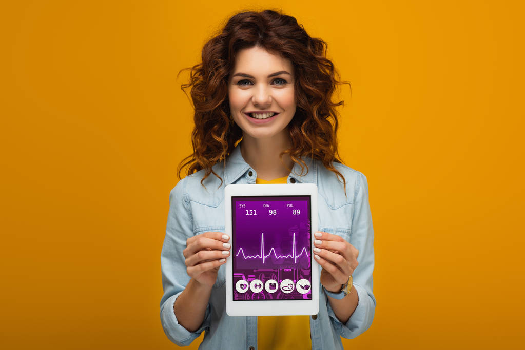 mulher ruiva alegre segurando tablet digital com aplicativo de e-saúde na tela enquanto está em pé na laranja
  - Foto, Imagem