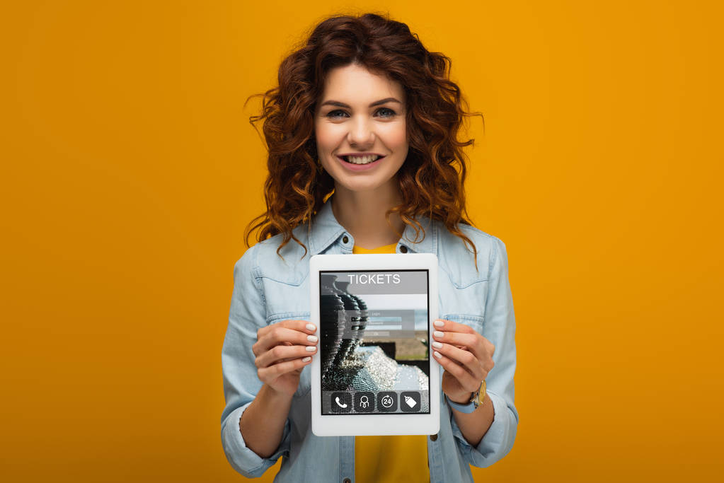 ευτυχισμένη κοκκινομάλλα γυναίκα κρατώντας ψηφιακό δισκίο με τα εισιτήρια app στην οθόνη, ενώ στέκεται στο πορτοκαλί  - Φωτογραφία, εικόνα