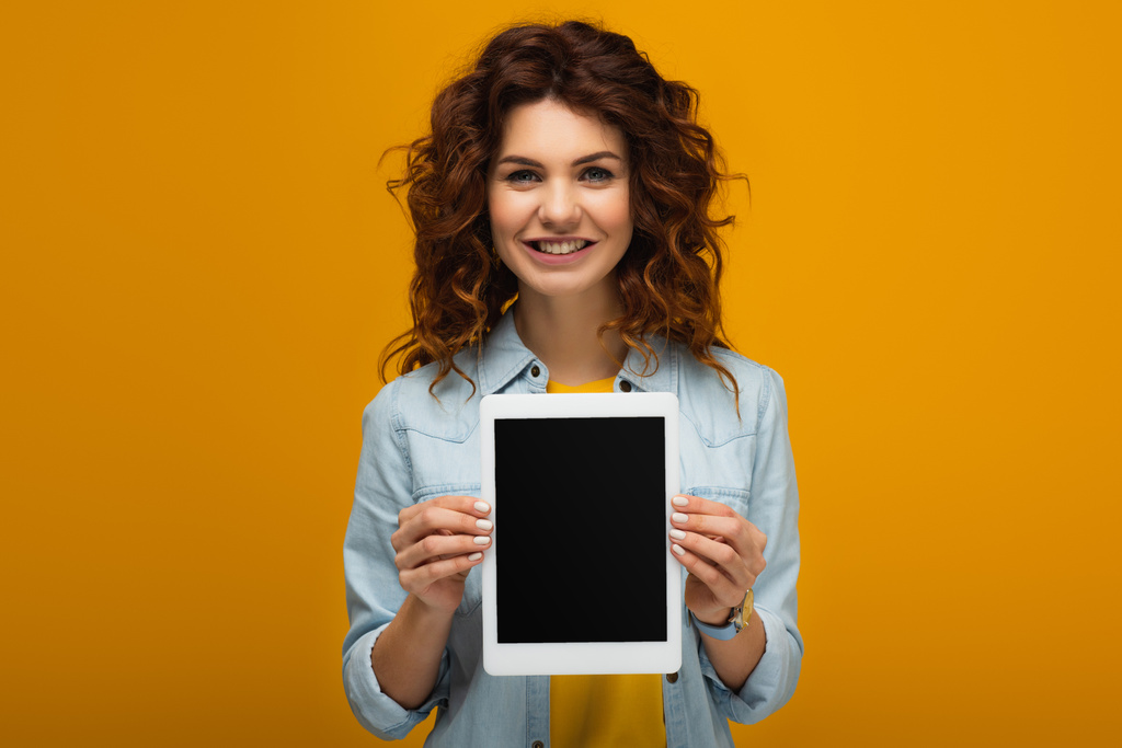 オレンジの上に立っている間、空白の画面でデジタルタブレットを保持する幸せな赤毛の女性  - 写真・画像