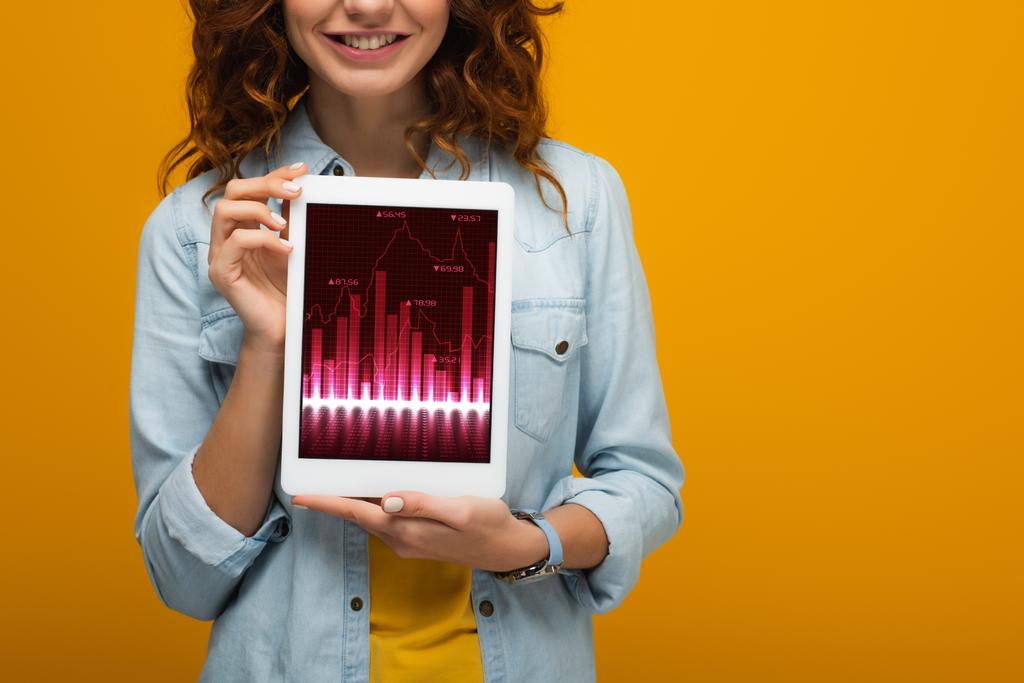turuncu izole ekranda grafikler ve grafikler ile dijital tablet tutan neşeli kıvırcık kız kırpılmış görünümü  - Fotoğraf, Görsel