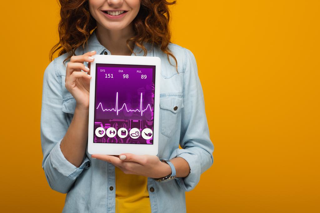 abgeschnittene Ansicht des fröhlichen lockigen Mädchens mit digitalem Tablet mit E-Health-App auf dem Bildschirm isoliert auf orange  - Foto, Bild