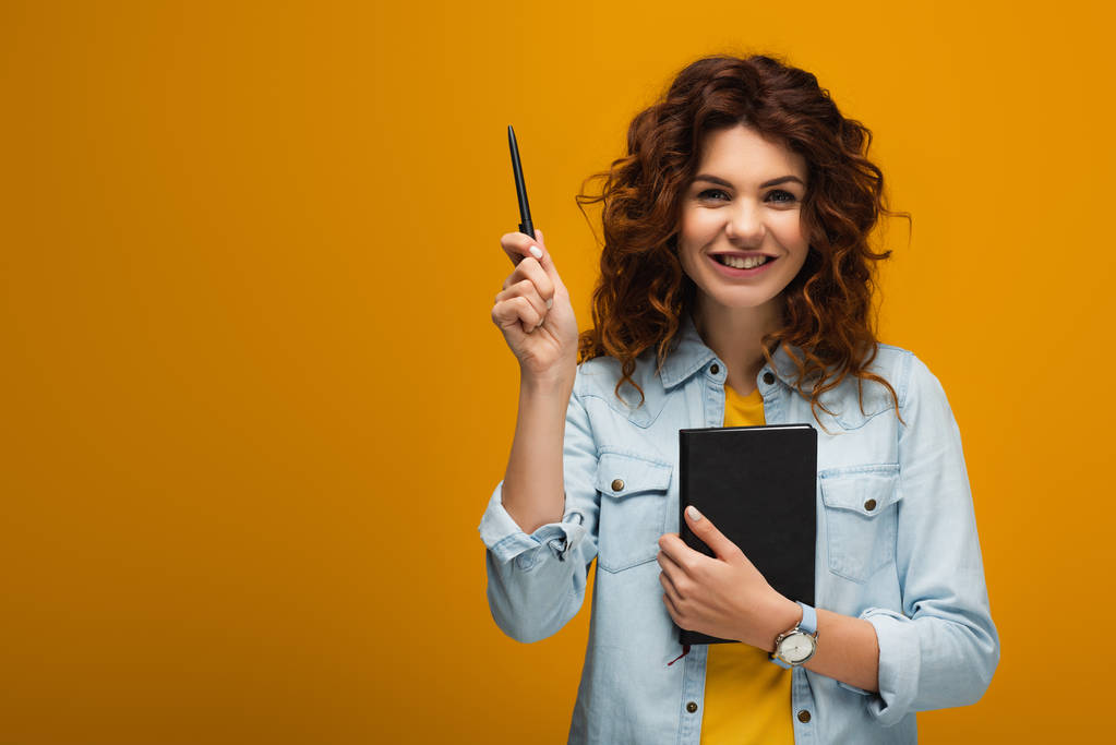 Gelukkig roodharige jonge vrouw houden notebook en pen op oranje  - Foto, afbeelding