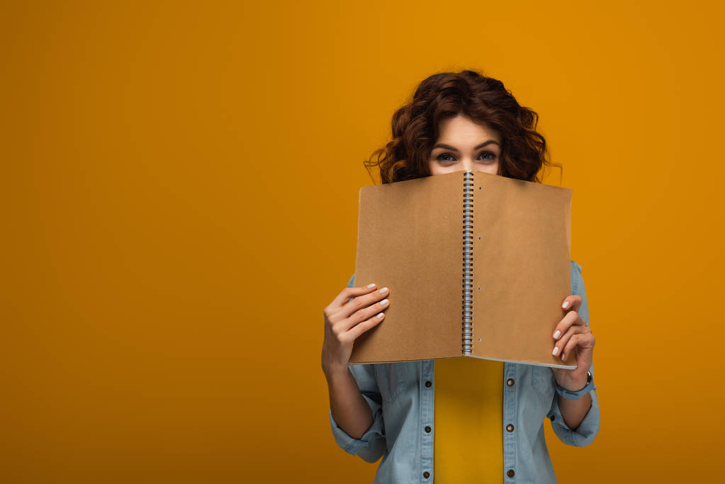 young curly redhead woman covering face with notebook on orange  - Photo, Image