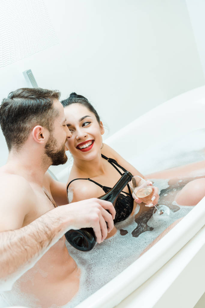 весёлый бородатый мужчина держит бутылку возле бокала шампанского счастливой женщины в ванной
  - Фото, изображение