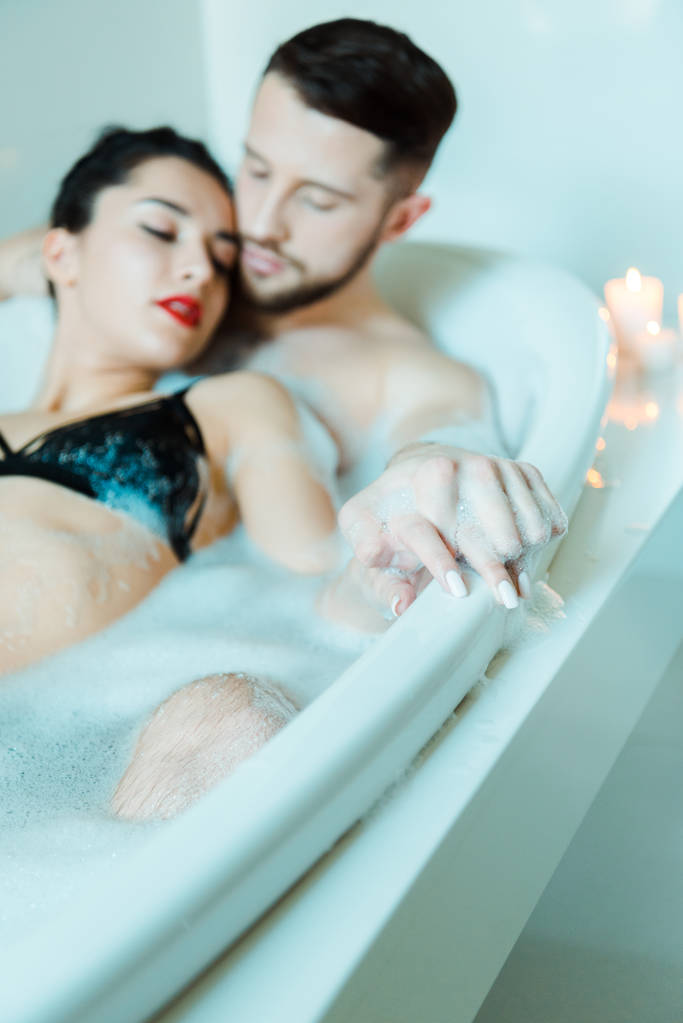 foco seletivo de homem barbudo e mulher morena atraente de mãos dadas enquanto deitado na banheira com espuma de banho
  - Foto, Imagem