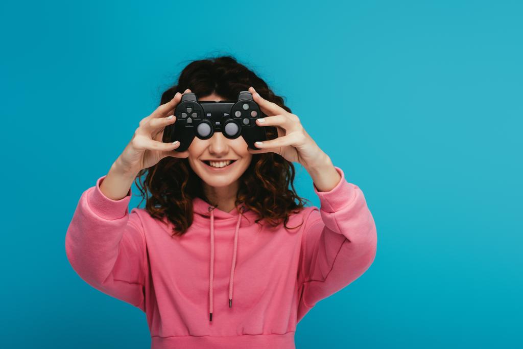 cheerful curly redhead girl covering face with joystick on blue - Photo, Image
