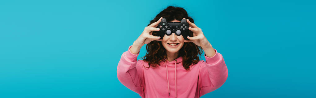 panoramatický záběr šťastné kudrnaté dívky zakrývající tvář pákovým ovladačem na modrém - Fotografie, Obrázek