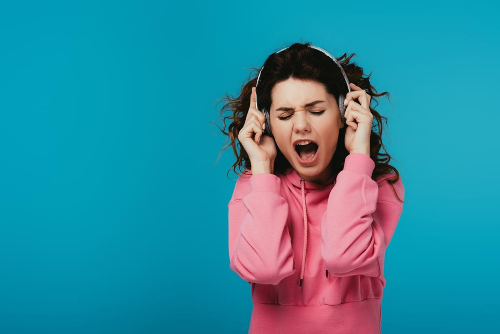 mulher encaracolado atraente cantando enquanto ouve música em fones de ouvido em azul
  - Foto, Imagem