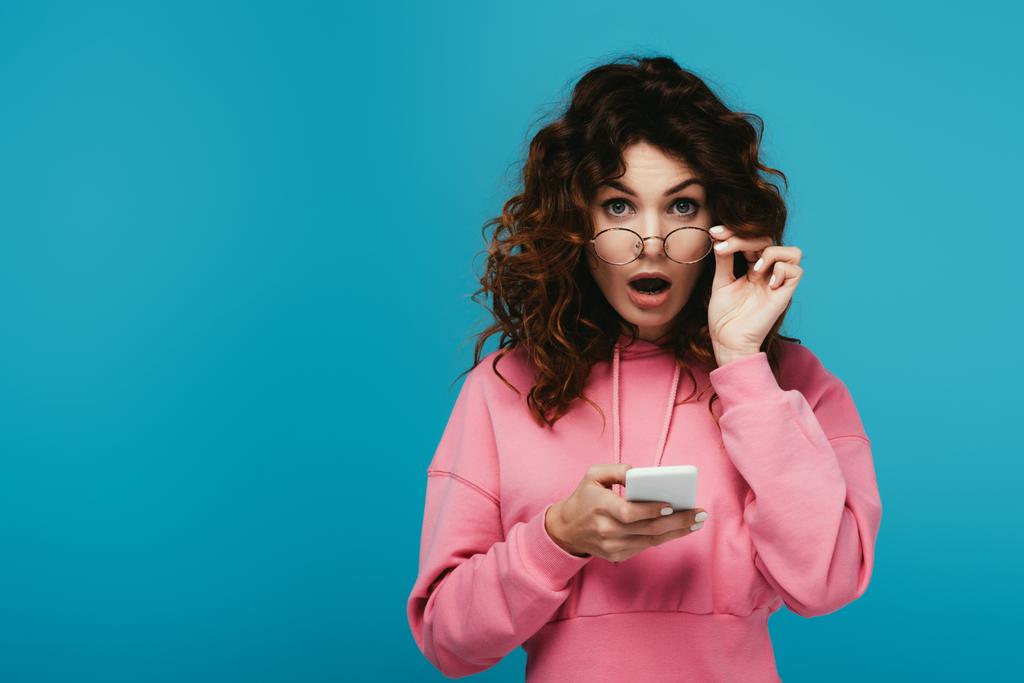chica rizada sorprendida tocando gafas y sosteniendo el teléfono inteligente en azul
 - Foto, Imagen