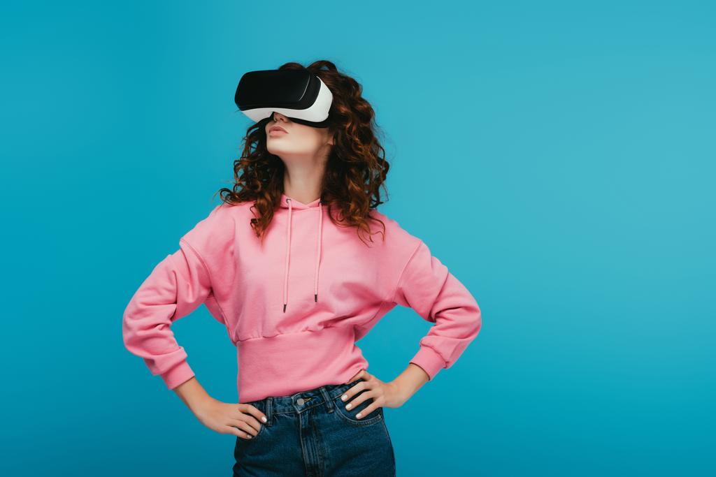 kręcone Dziewczyna nosi zestaw słuchawkowy wirtualnej rzeczywistości stojąc z rękami na biodrach na niebiesko  - Zdjęcie, obraz