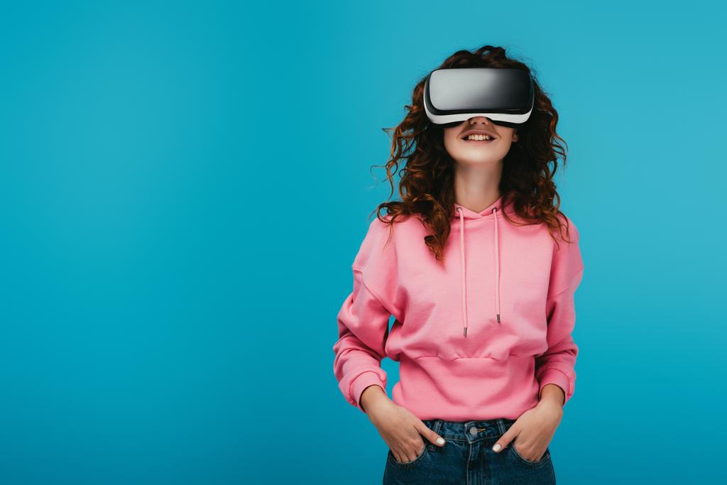 curly girl wearing virtual reality headset while standing with hands in pockets on blue  - Photo, Image