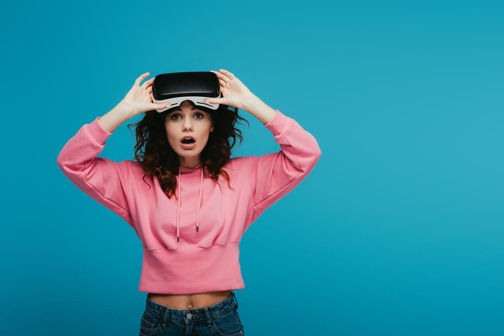 chica rizada sorprendida con auriculares de realidad virtual en azul
  - Foto, Imagen