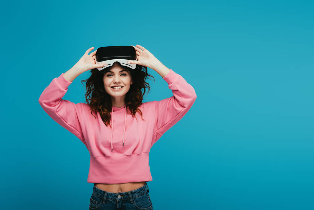 cheerful curly girl wearing virtual reality headset and smiling on blue  - Photo, Image