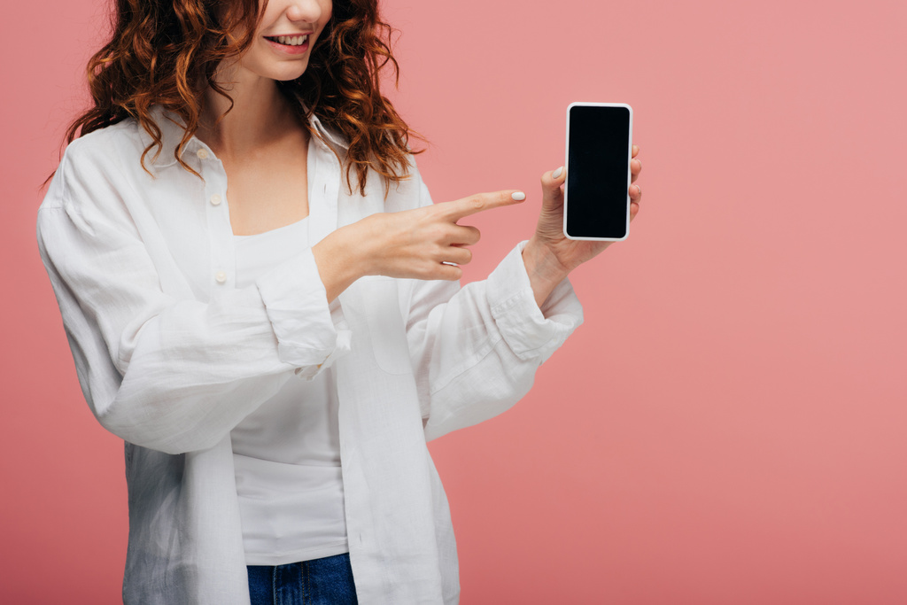 Ausgeschnittene Ansicht eines fröhlichen Mädchens, das mit dem Finger auf ein Smartphone mit leerem Bildschirm auf rosa zeigt - Foto, Bild