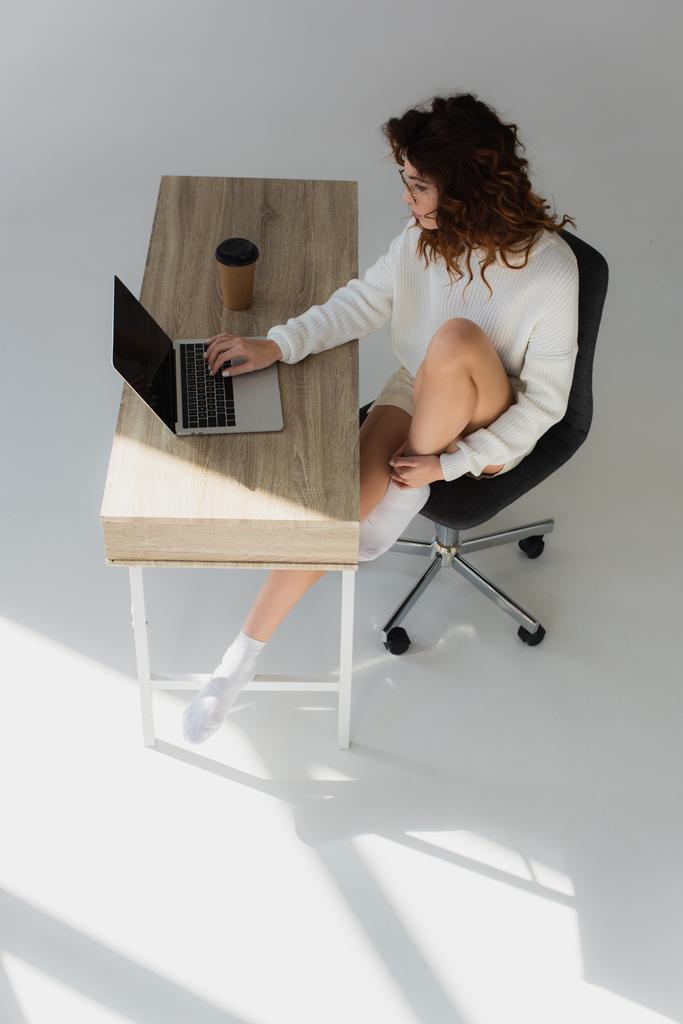 над головой вид молодой женщины в очках, сидящей и использующей ноутбук рядом с бумажной чашкой на сером
  - Фото, изображение