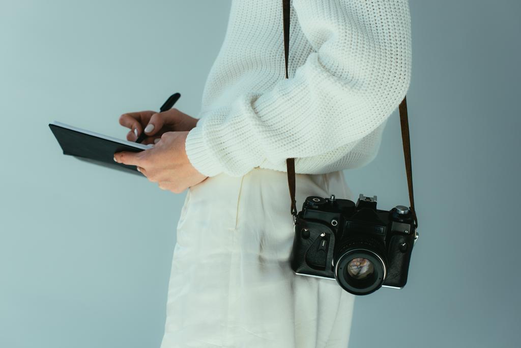 Ausgeschnittene Ansicht einer jungen Frau, die im Stehen mit Digitalkamera in ein Notizbuch schreibt  - Foto, Bild