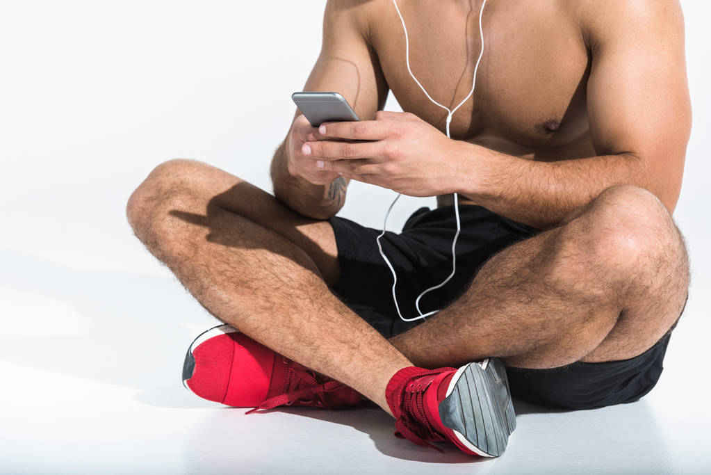ostříhané zobrazení sportivního muže se smartphone, který sedí na podlaze s zkřížené nohy na bílém - Fotografie, Obrázek
