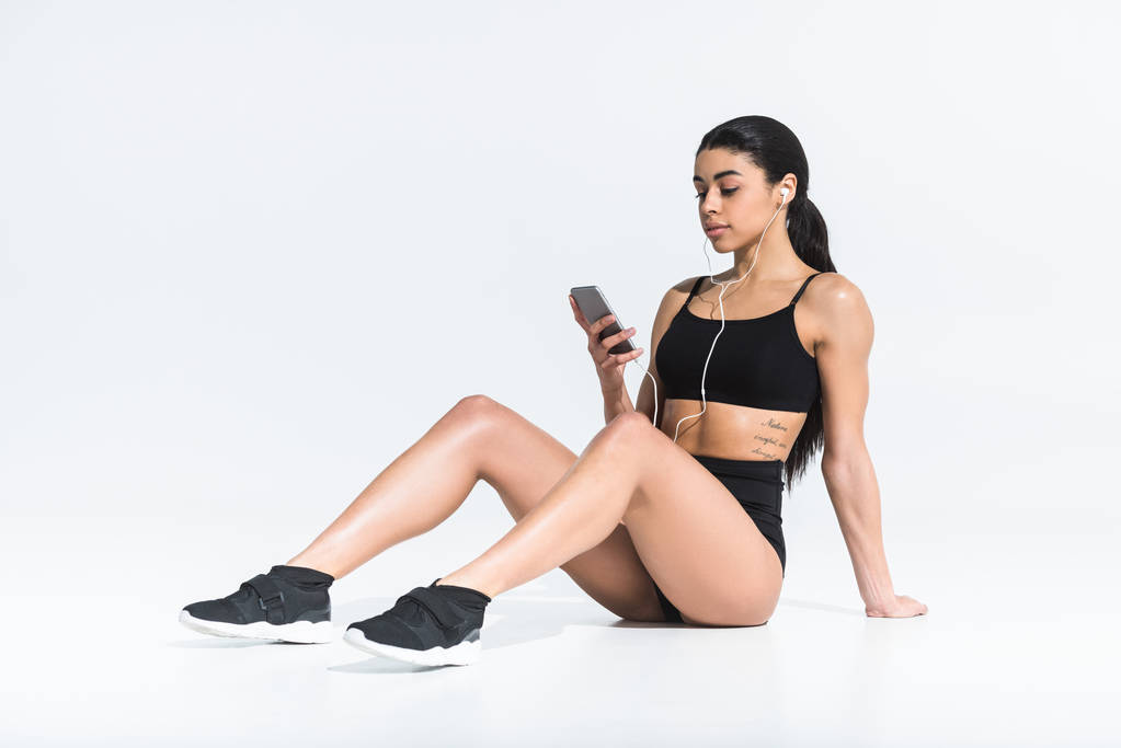 attractive african american girl in sportswear sitting on floor and listening music in earphones on white - Photo, Image