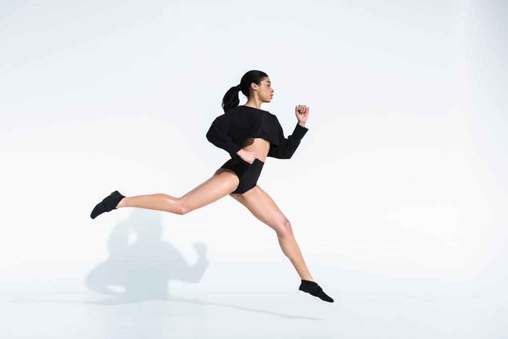 vista lateral de la chica afroamericana deportiva en ropa deportiva negra y zapatillas de deporte corriendo en blanco
 - Foto, Imagen