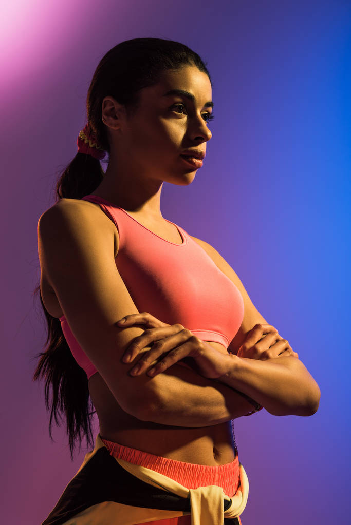 chica afroamericana deportiva en sujetador deportivo rosa posando con las manos cruzadas sobre fondo de degradado púrpura y azul
 - Foto, Imagen