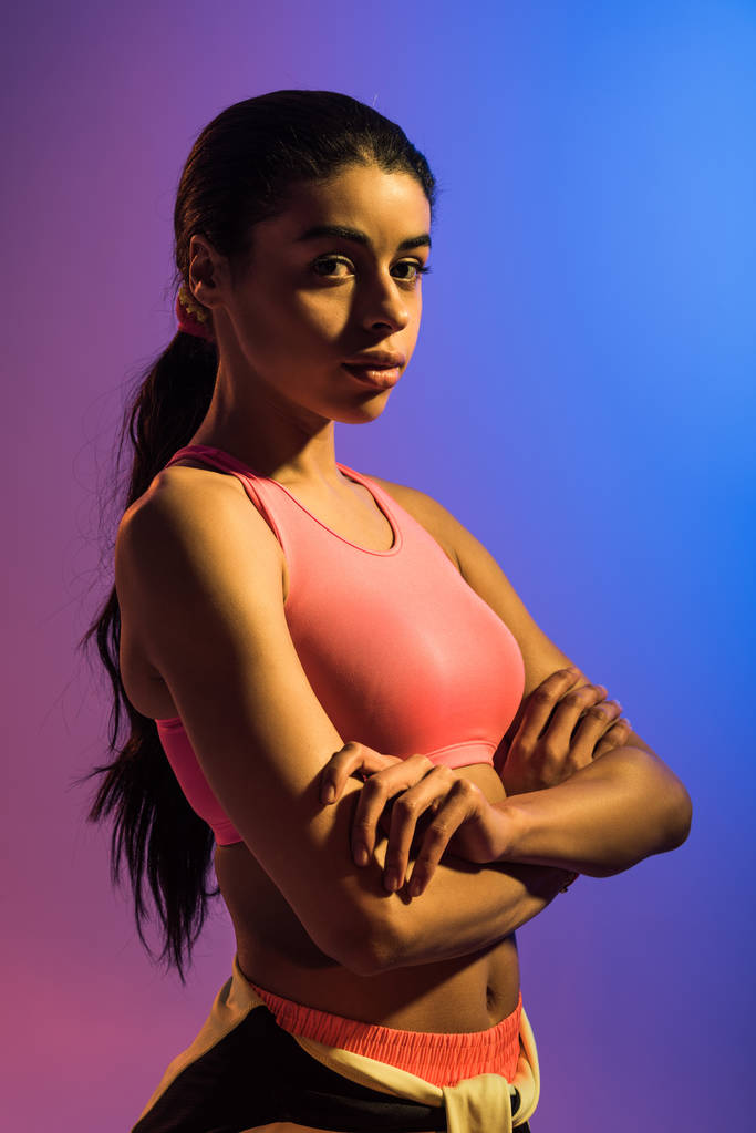 piękna Sportive African American Girl pozowanie w aparacie z skrzyżowanymi ramionami na fioletowym i niebieskim tle gradientu - Zdjęcie, obraz