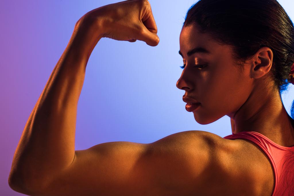 mooie atletische African American Girl demonstreren biceps op paarse en blauwe gradiënt achtergrond - Foto, afbeelding