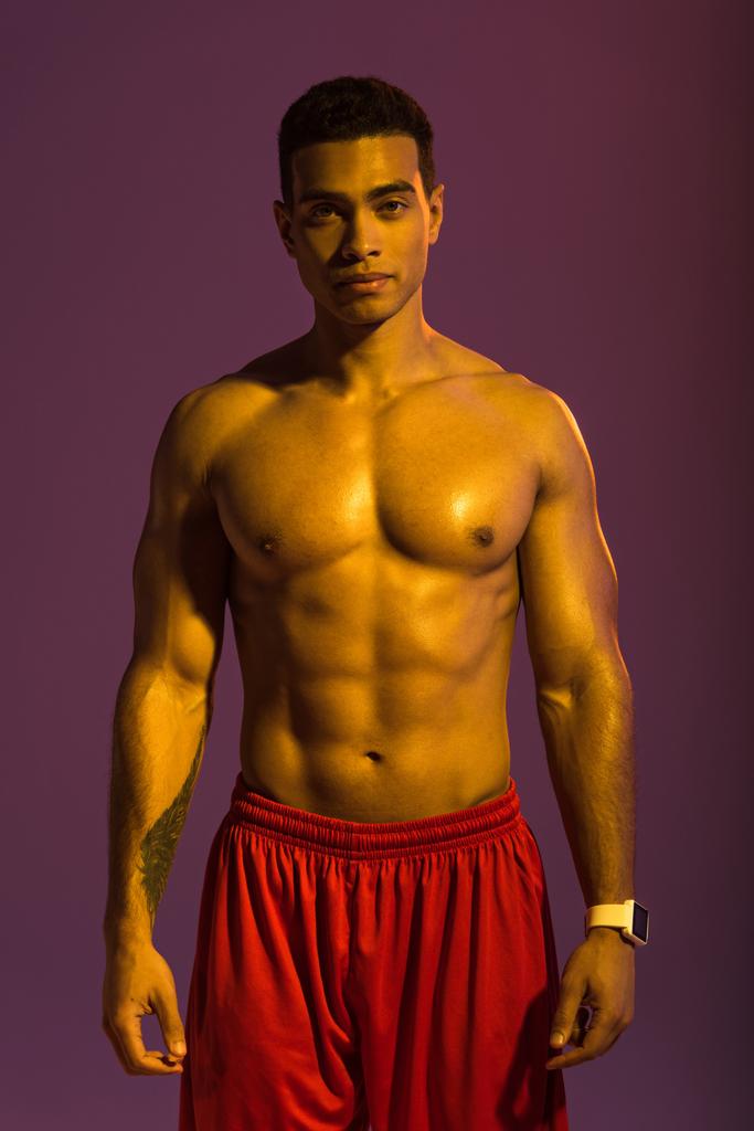 guapo deportivo mixto hombre de raza en reloj inteligente y pantalones cortos rojos mirando a la cámara sobre fondo púrpura
 - Foto, imagen