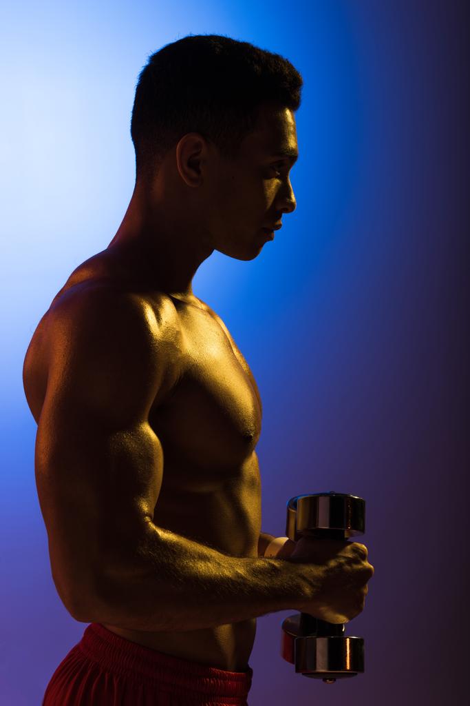 vista laterale di atletico uomo razza mista con manubri su sfondo sfumato blu e viola scuro
 - Foto, immagini