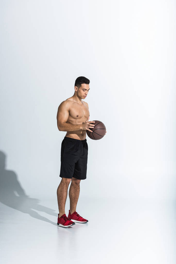 όμορφος/η γυμνός μικτός άνθρωπος του αγώνα με μαύρο σορτς και κόκκινα αθλητικά παπούτσια κρατώντας μπάλα σε λευκό - Φωτογραφία, εικόνα