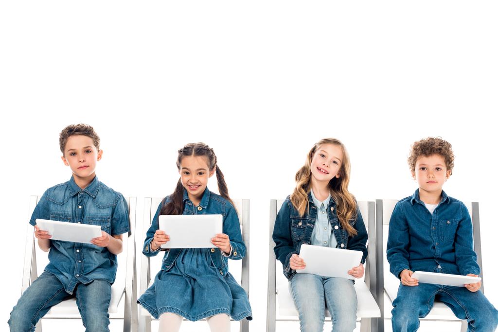 cztery uśmiechnięte dzieci w dżinsowych ubraniach siedzących na krzeseł i wykorzystujących cyfrowe tablety na białym tle - Zdjęcie, obraz