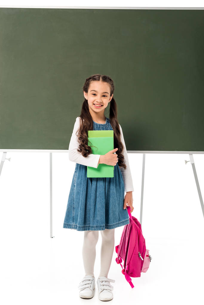pohled na usměvavou školku s batohu a knihami stojící poblíž černé tabule izolovaná na bílém - Fotografie, Obrázek