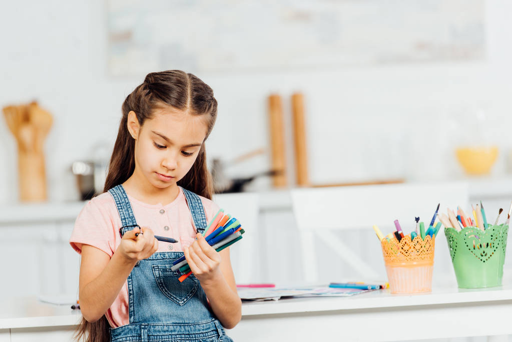 cute kid holding colorful felt pens and pen while standing near table at home  - Photo, Image