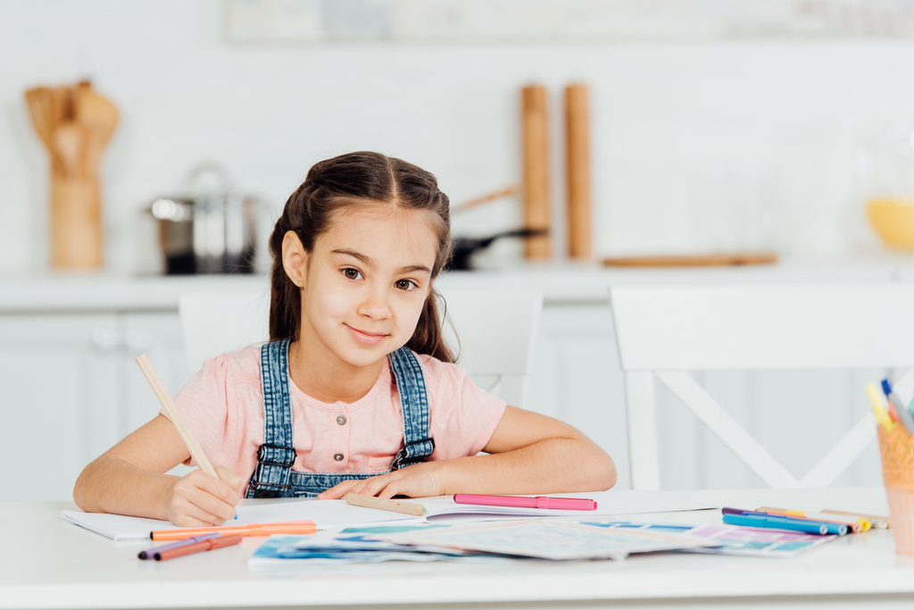 χαριτωμένο παιδί κοιτάζοντας την κάμερα, κρατώντας το χρώμα μολύβι κοντά στο χαρτί στο σπίτι  - Φωτογραφία, εικόνα