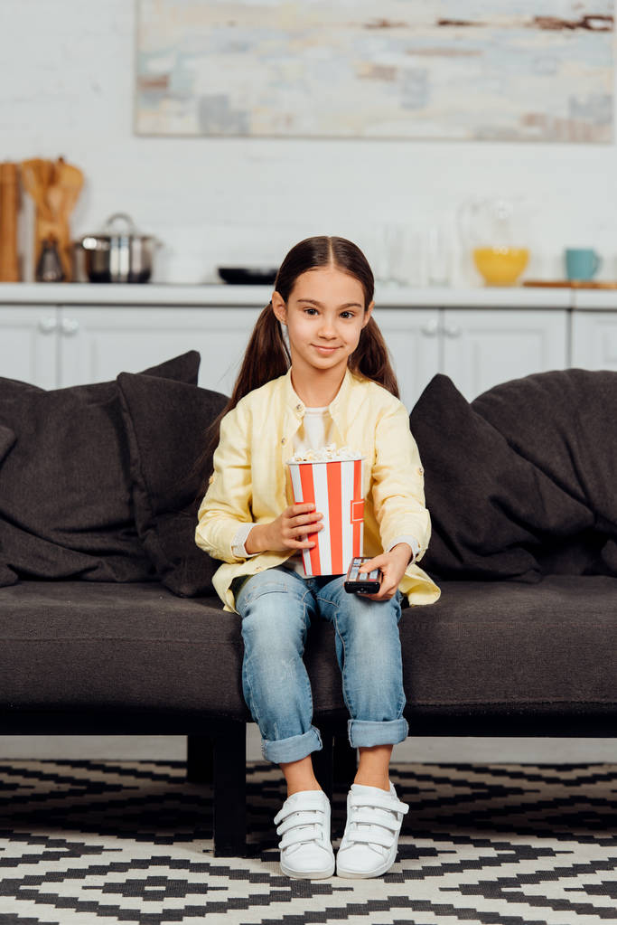 szczęśliwy Kid trzymając pilota zdalnego sterowania siedząc na kanapie z wiadrem popcorn  - Zdjęcie, obraz
