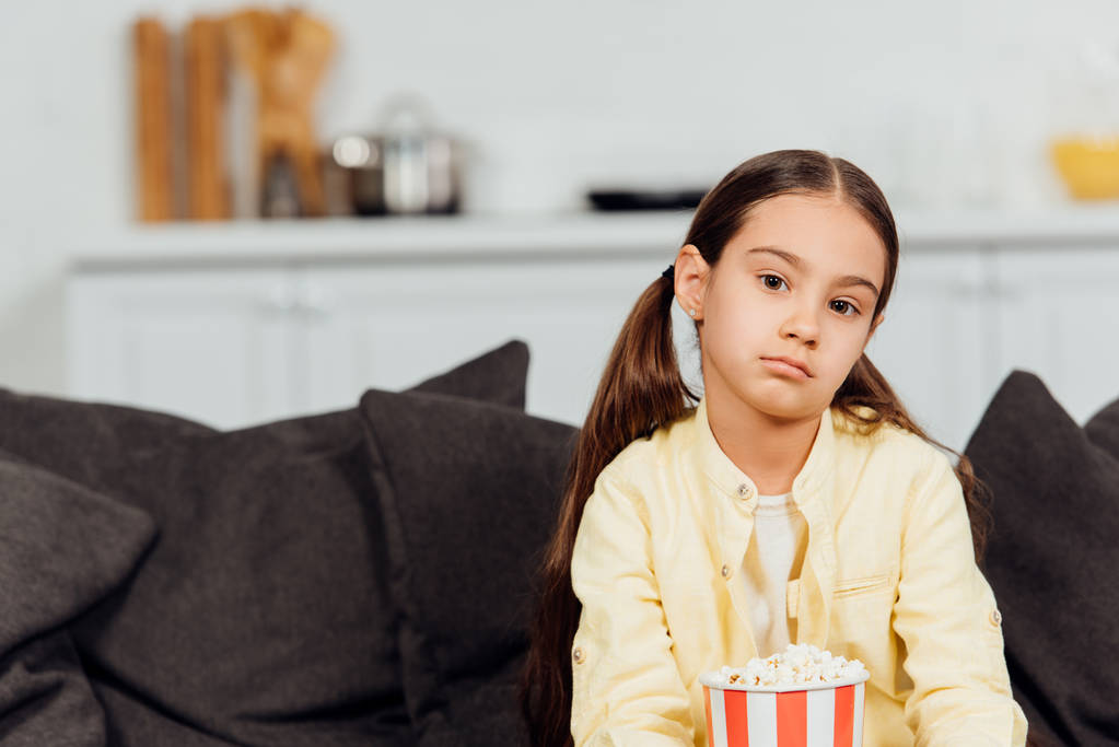 sad and cute kid looking at camera while sitting with bucket of popcorn  - Photo, Image