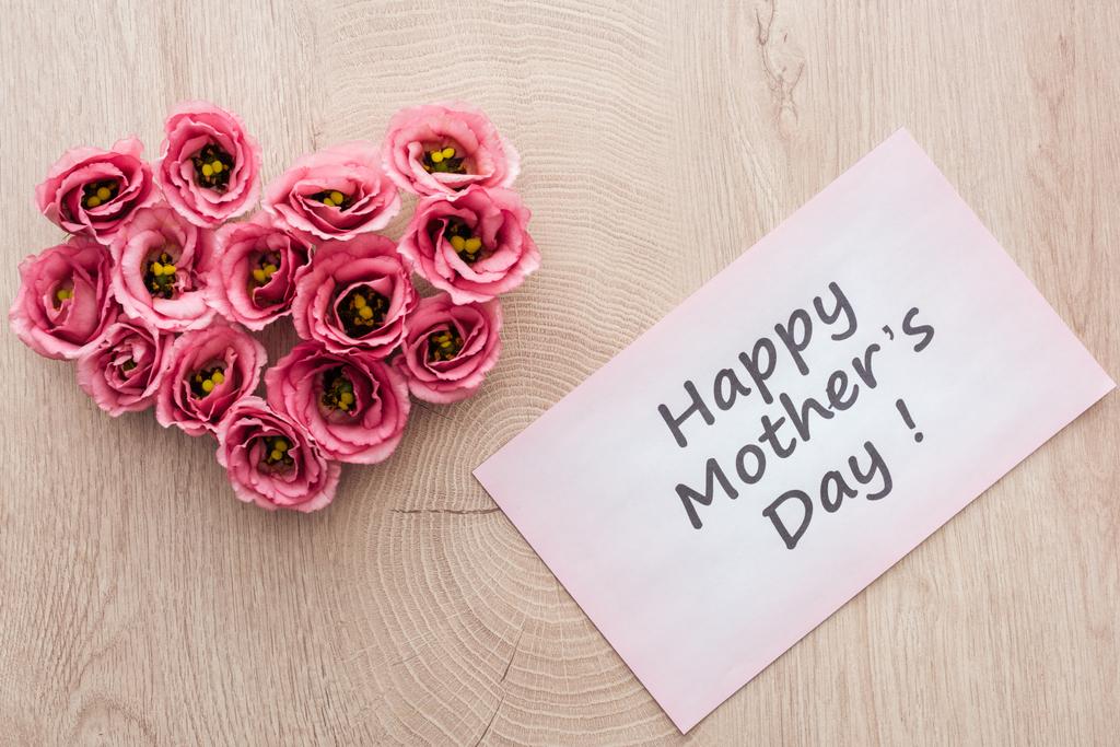 Herzschild aus Eustoma-Blumen und Grußkarte mit fröhlichem Muttertagsaufdruck auf Holztisch - Foto, Bild