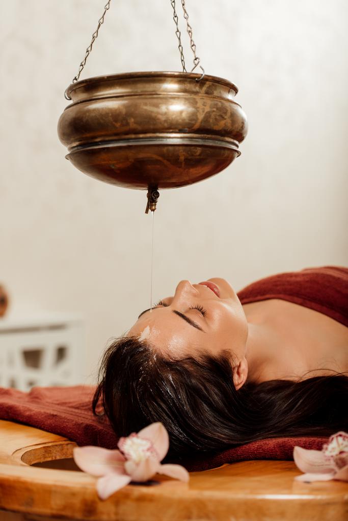 mujer joven relajada acostada bajo el vaso shirodhara durante el procedimiento ayurvédico
 - Foto, Imagen