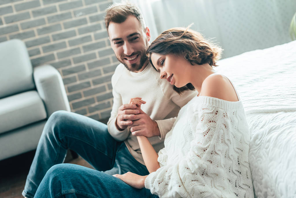 glücklicher Mann hält Händchen mit attraktiver Freundin, während er zu Hause auf dem Boden sitzt  - Foto, Bild
