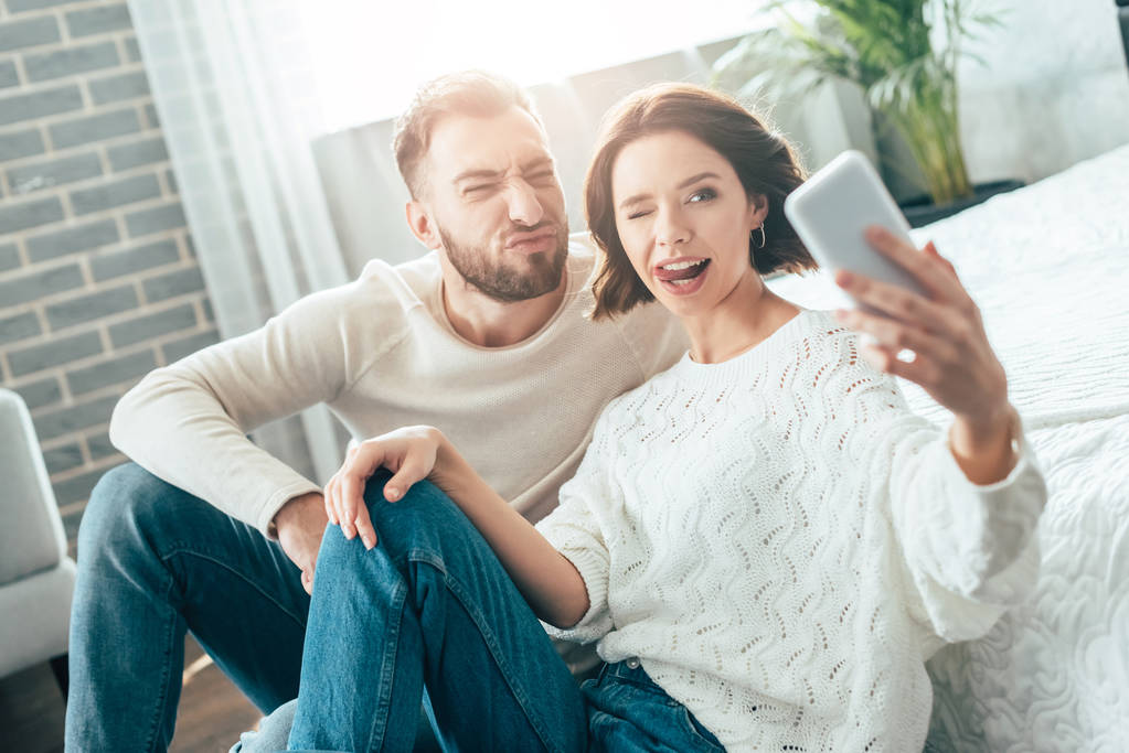 χαρούμενη γυναίκα που δείχνει τη γλώσσα, ενώ παίρνει selfie με μούσι άντρα στο σπίτι  - Φωτογραφία, εικόνα