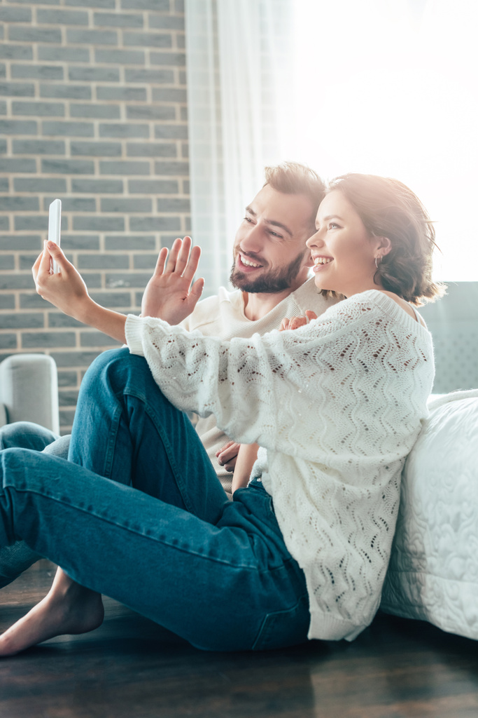 χαρούμενη γυναίκα κουνώντας το χέρι, ενώ παίρνει selfie με τον άνθρωπο στο σπίτι  - Φωτογραφία, εικόνα