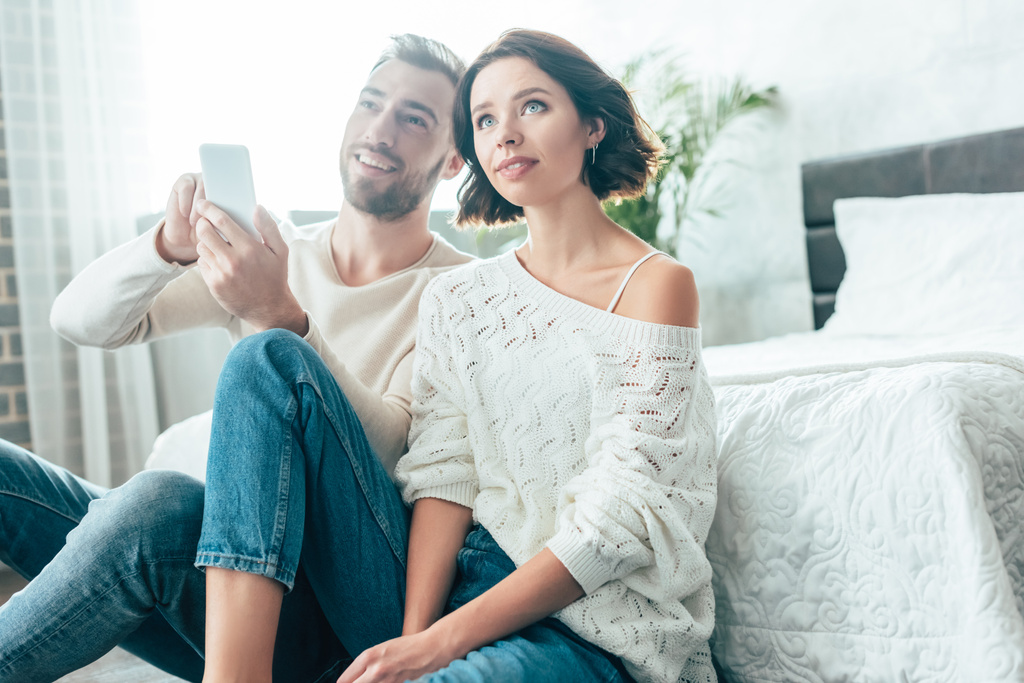 šťastný muž držící smartphone blízko zasněný bruneta doma  - Fotografie, Obrázek