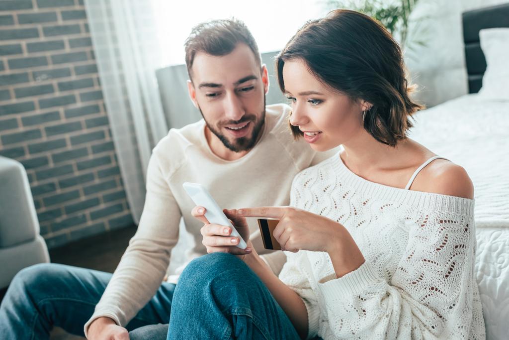 attraktive Frau zeigt mit dem Finger auf Smartphone und hält Kreditkarte in der Nähe glücklichen Mannes  - Foto, Bild