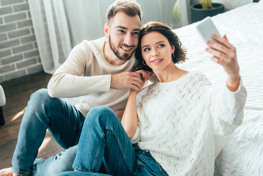 selektiver Fokus attraktiver Frau und gutaussehender Mann, die sich Händchen halten, während sie zu Hause ein Selfie machen  - Foto, Bild